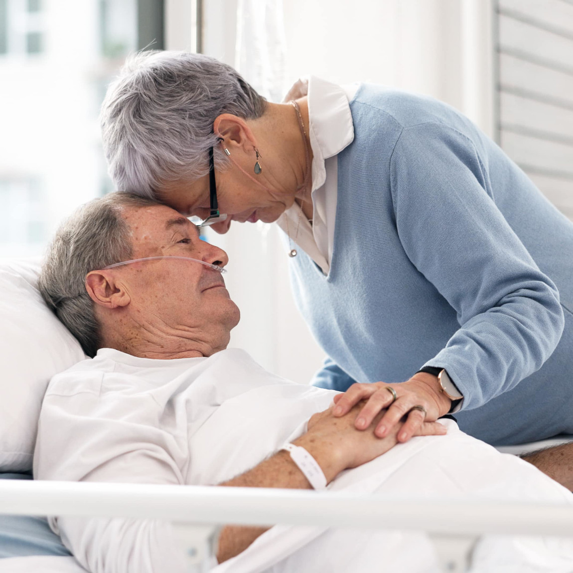 Eine ältere Frau beugt sich liebevoll über einen älteren Mann, der im Krankenhausbett liegt, symbolisiert emotionale Unterstützung in einer schwierigen Zeit, möglicherweise im Rahmen der Vorsorgeplanung