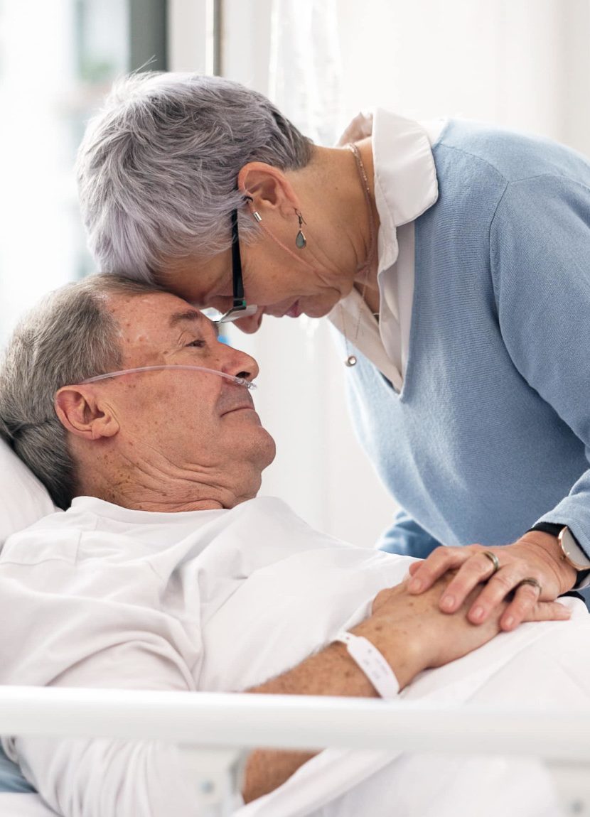Eine ältere Frau beugt sich liebevoll über einen älteren Mann, der im Krankenhausbett liegt, symbolisiert emotionale Unterstützung in einer schwierigen Zeit, möglicherweise im Rahmen der Vorsorgeplanung